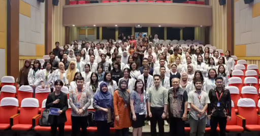 A full lecture hall of young participants in rows of red seats.